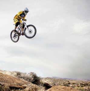 A cyclist performs a daring jump off a rocky cliff under a cloudy sky.