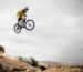 A cyclist performs a daring jump off a rocky cliff under a cloudy sky.